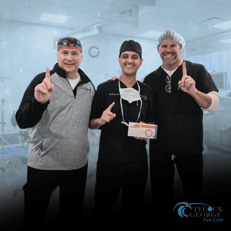Three smiling men stand together in a medical setting, all holding up one finger to indicate “number one.” The man in the center, wearing black scrubs and a surgical cap, holds a box labeled “Preloaded IOL.” The other two men wear black and gray tops and surgical head coverings. The Tylock George Eye Care logo appears in the lower right corner.